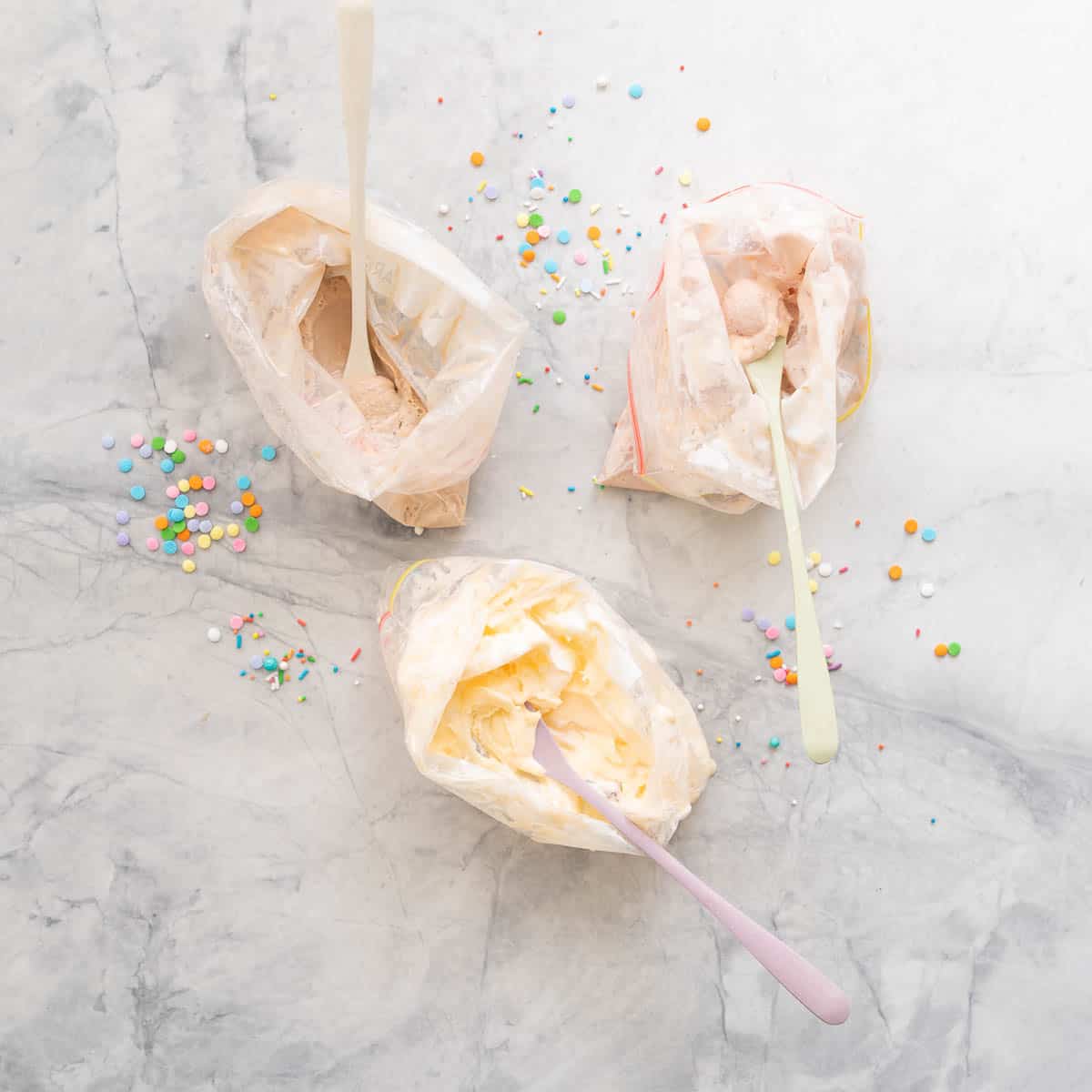 Three ziplock bags of ice cream with spoons on a marble bench scattered with sprinkles.