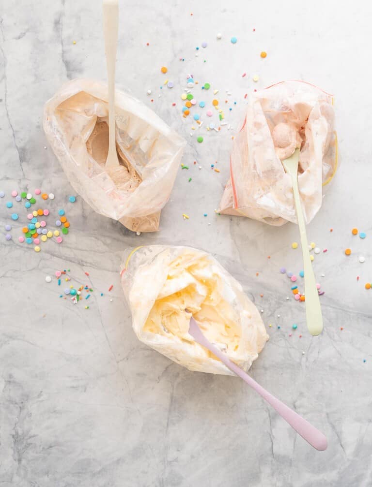 Three ziplock bags of ice cream with spoons on a marble bench scattered with sprinkles.