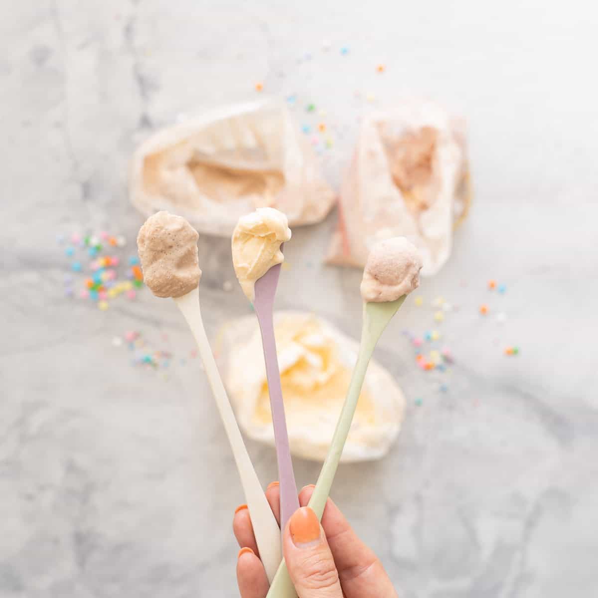 Three spoonfuls of different flavoured ice cream (chocolate, ice cream and strawberry) being held above bags if ice cream.