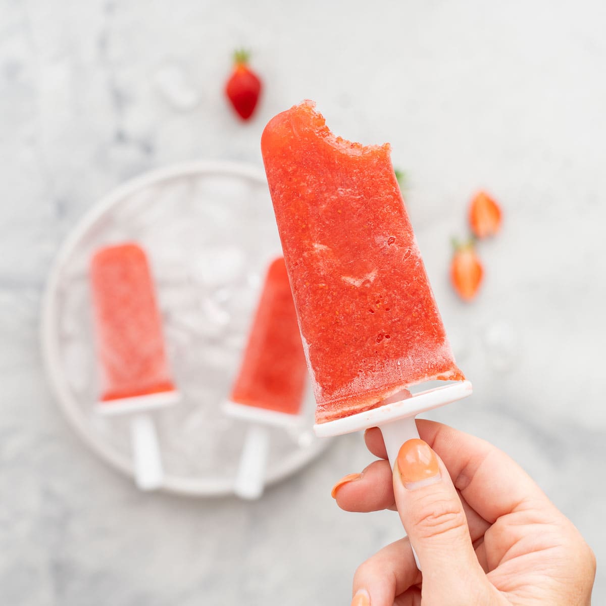A strawberry popsicle with one bite taken out of it being held up to the camera.