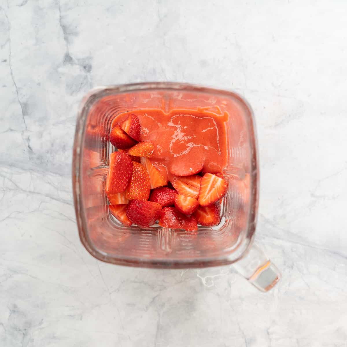 A glass blender jug with sliced strawberries and strawberry puree in it