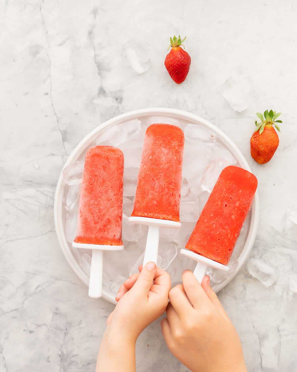 Childrens hands reaching to pick up popsicles from a bowl filled with ice.
