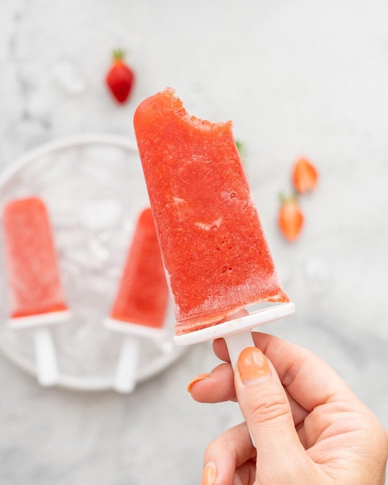 A strawberry popsicle with one bite taken out of it being held up to the camera.