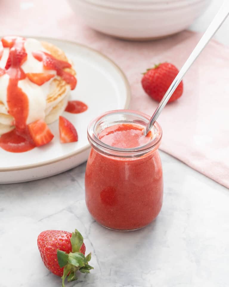 A small glass jar of strawberry puree in front of a plate of pancakes drizzled with the syrup.