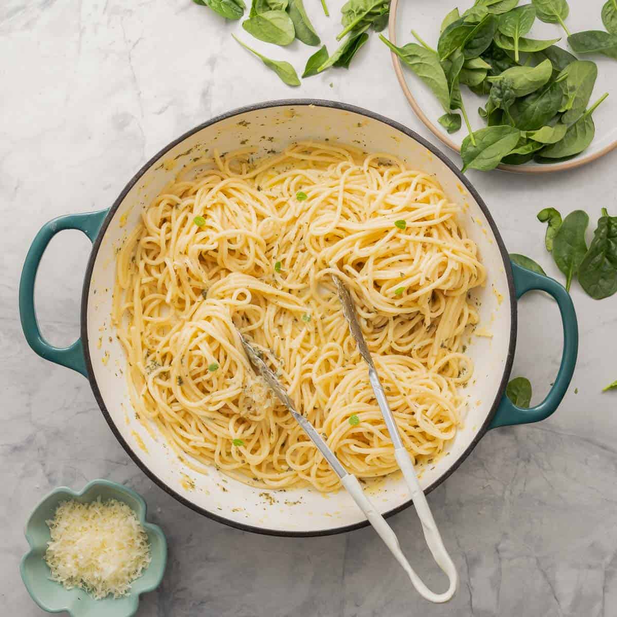  A large pan full of buttered noodles resting on the bench next to plate of spinach and a small ramekin of parmesan cheese. 