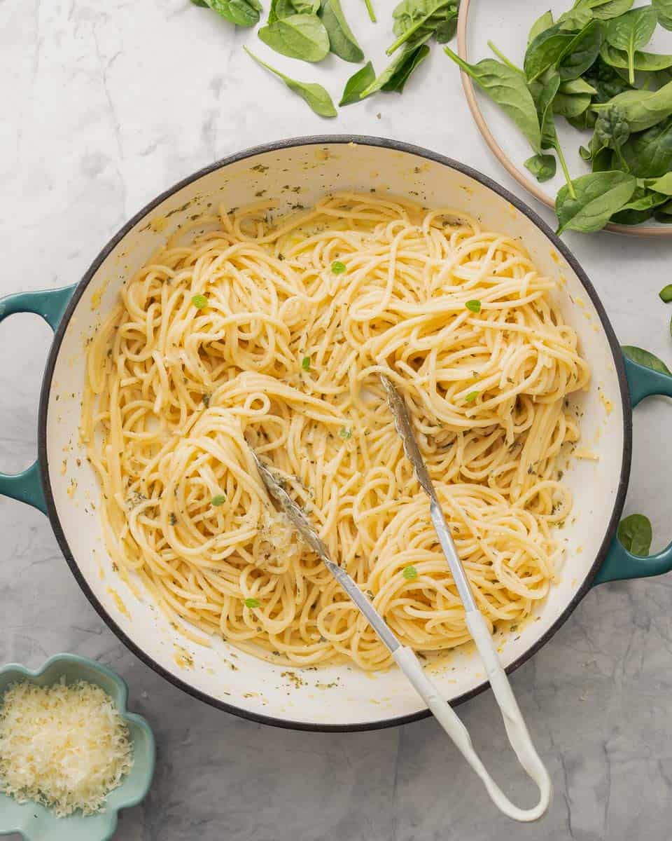 A large pan full of buttered noodles resting on the bench next to plate of spinach and a small ramekin of parmesan cheese.
