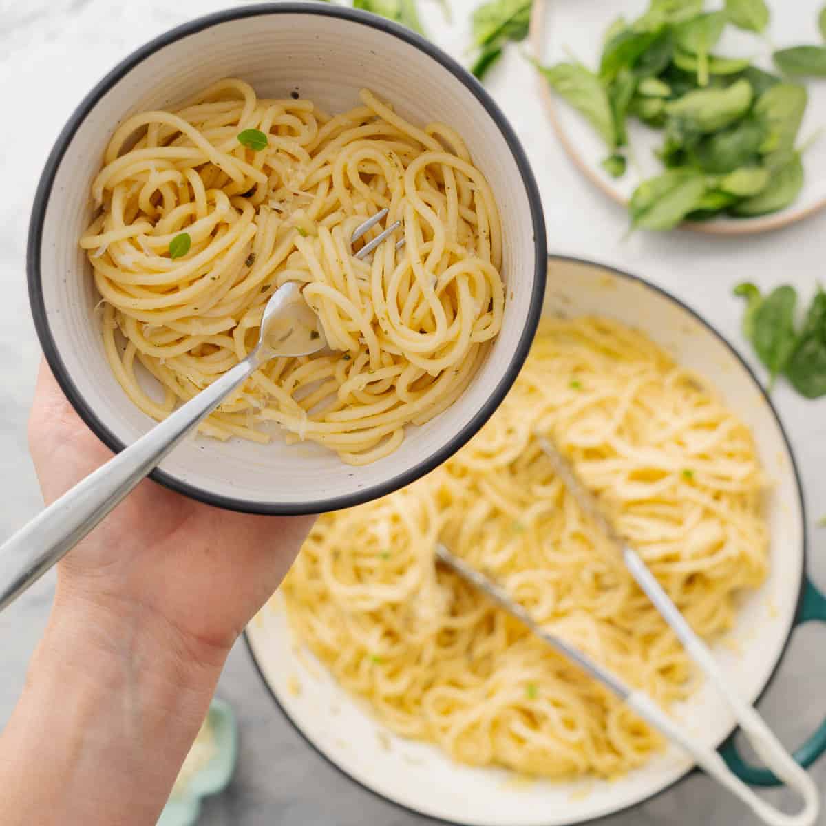 A large pan full of buttered noodles resting on the bench with a serving on a plate being held above it by a hand