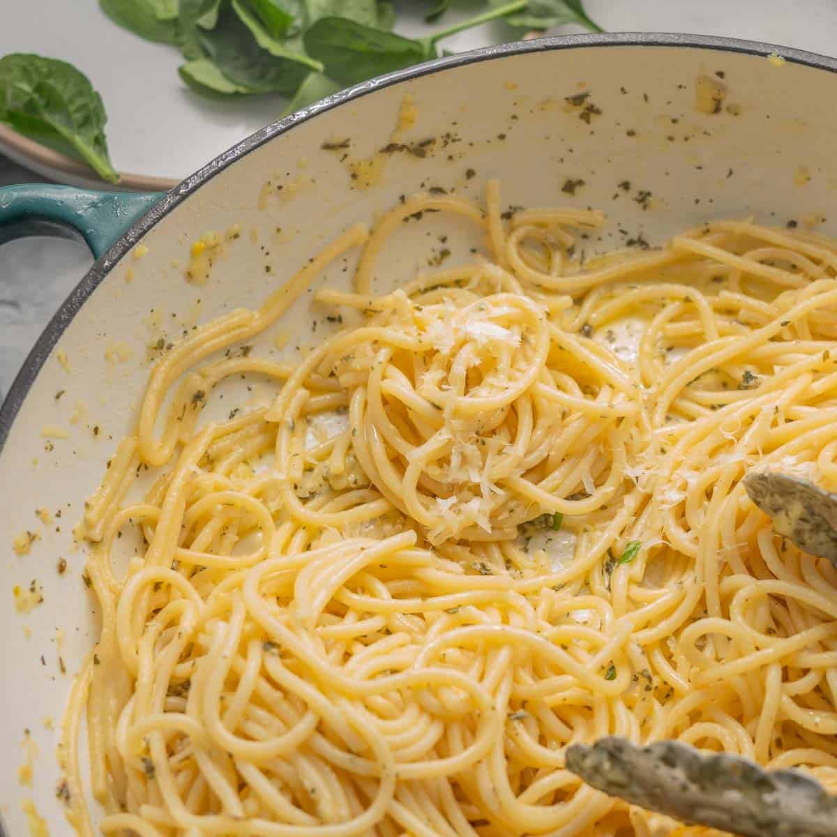 A large pan full of buttered noodles resting on the bench next to a scattering of spinach leaves