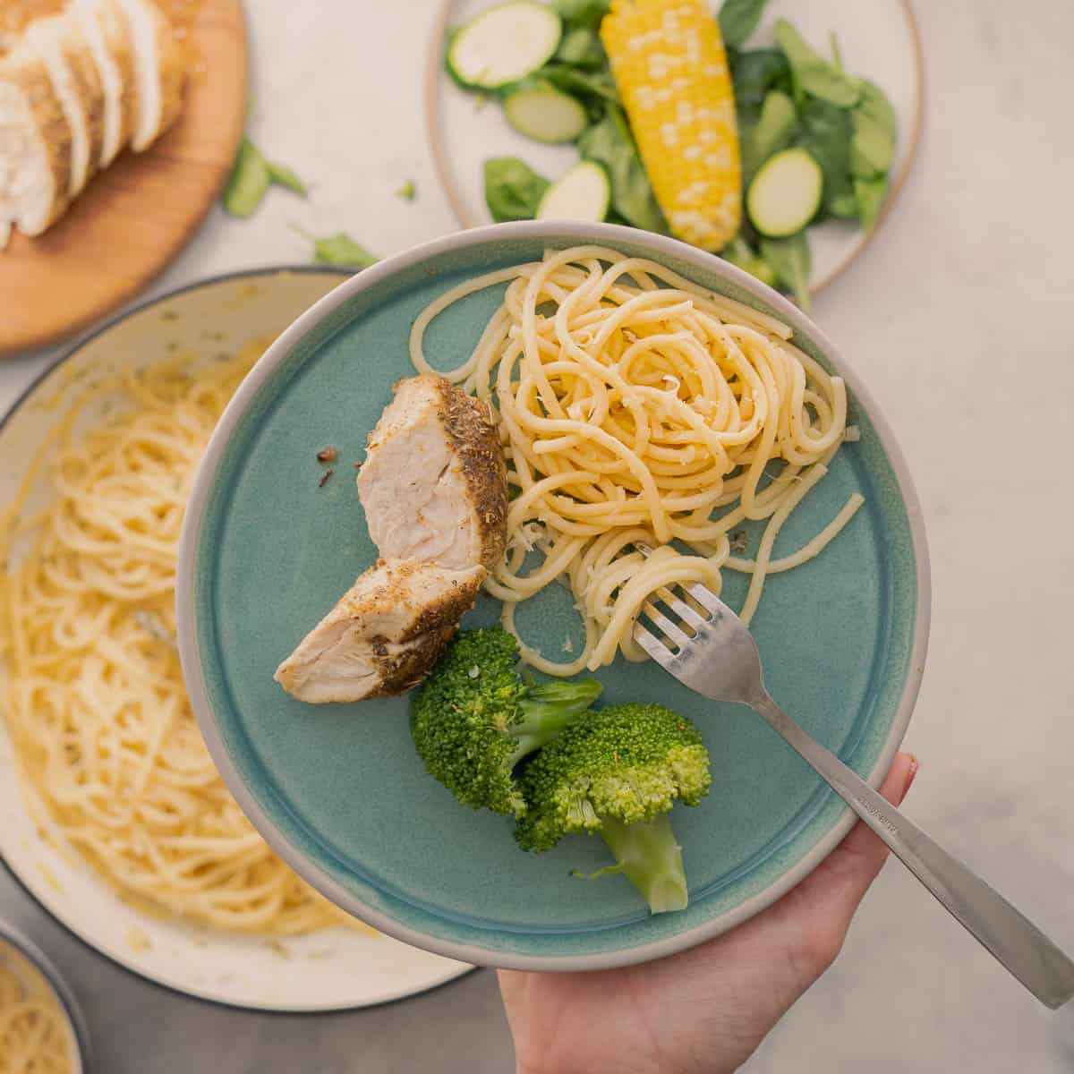 A large pan full of buttered noodles resting on the bench with a serving on a plate with cooked and sliced chicken and broccoli being held above it by a hand