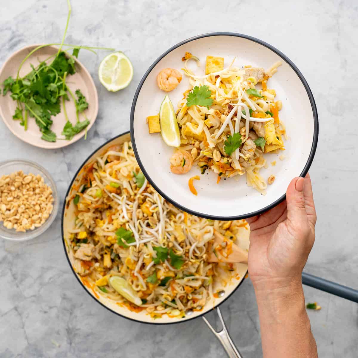 A white plate with black rim holding pad this, with bean sprouts, lime wedges and prawns visible above a large fry pan of pad thai.