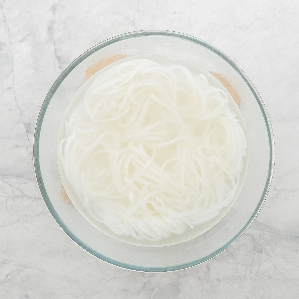 A glass bowl of rice stick noodles soaking in water.