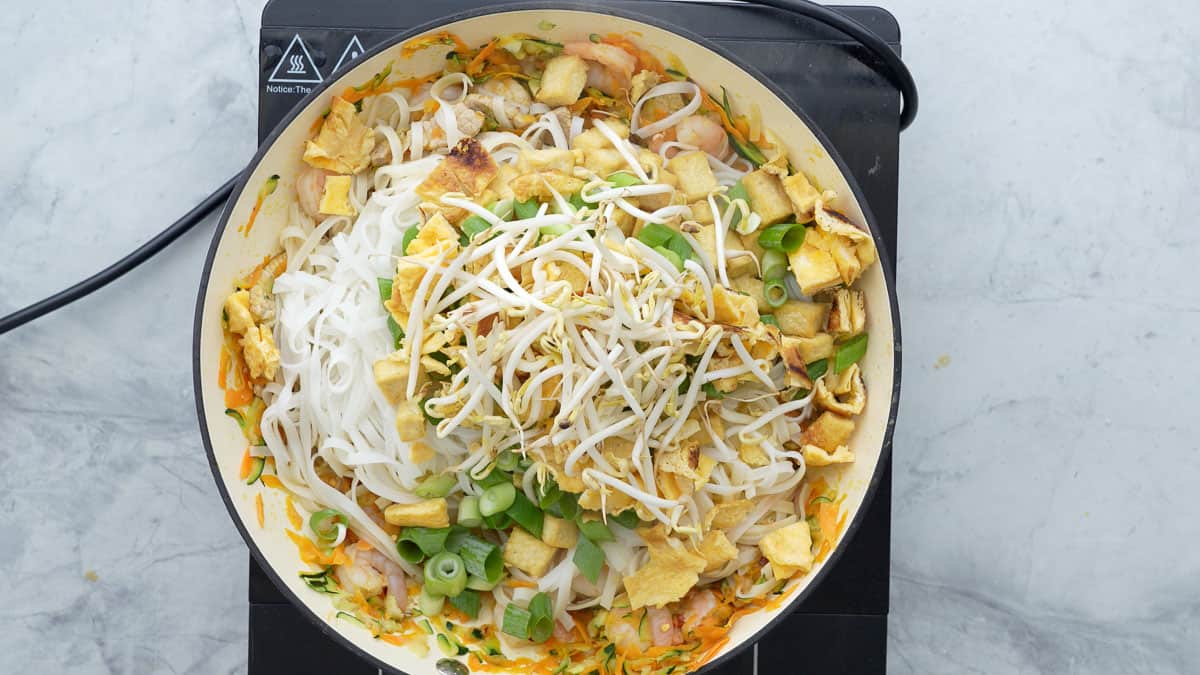 Noodles, eggs, tofu spring onions and pad thai sauce in a ceramic wok.