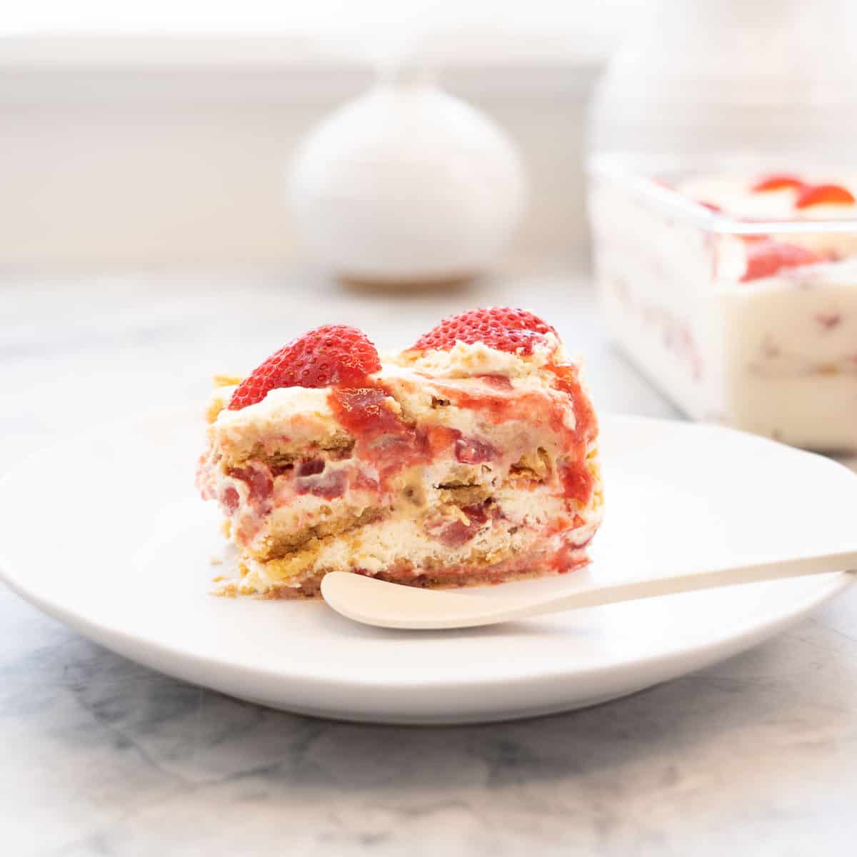 A piece of a strawberry shortcake ice box cake on a plate with a white bamboo spoon.