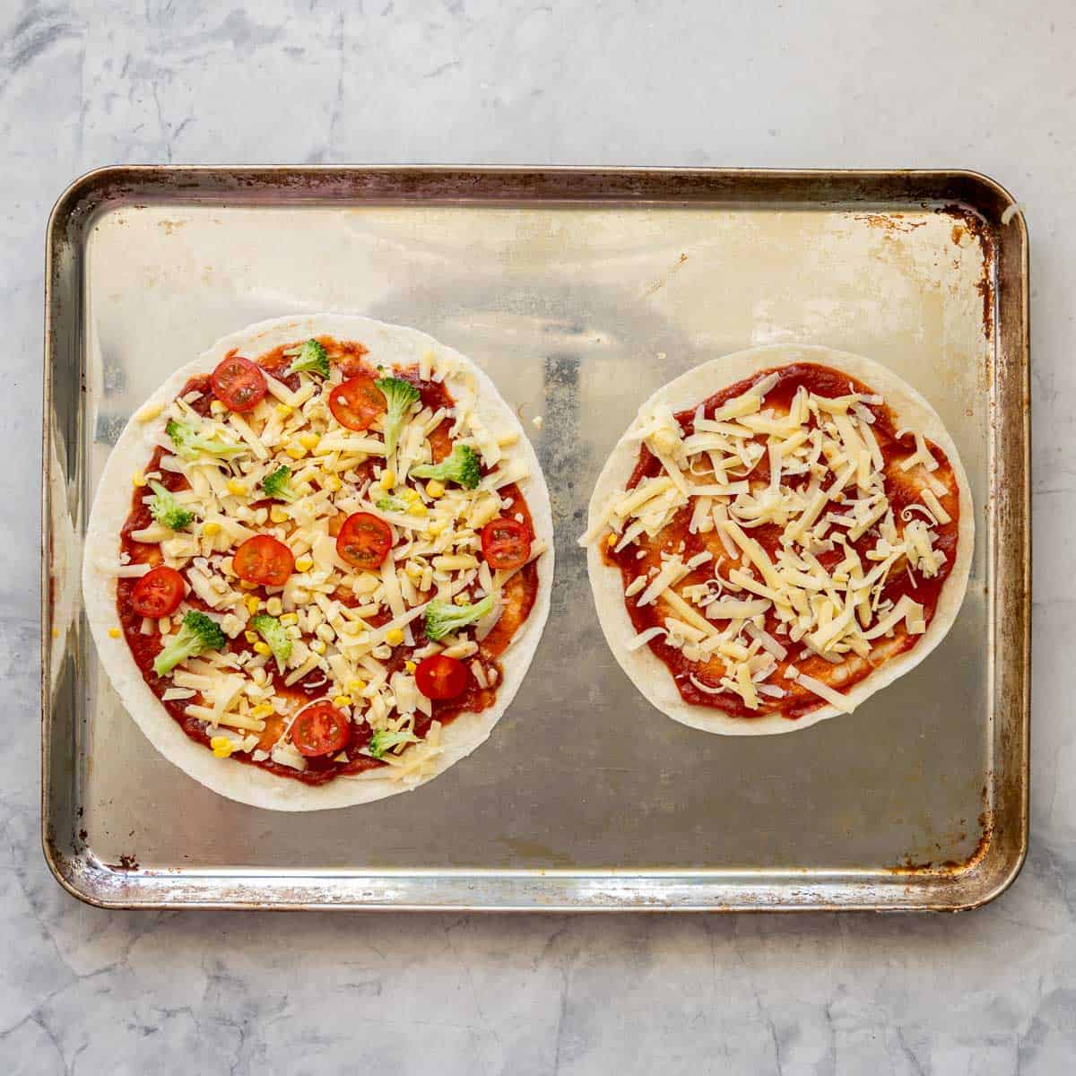 Two tortillas on a baking tray, spread with pizza sauce and toppings and cheese