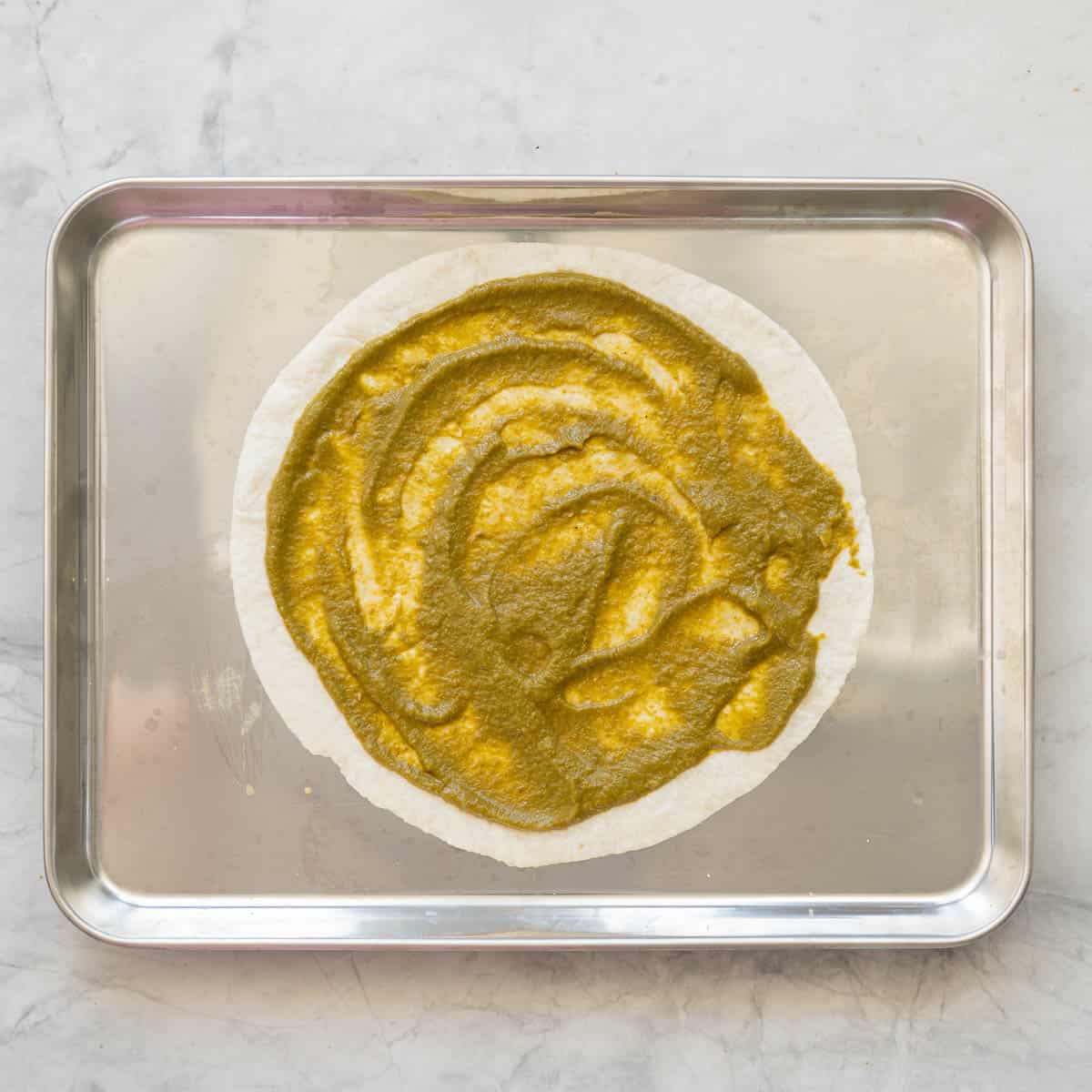 An oven tray resting on the bench with a tortilla spread with vegetable pizza sauce on top of it.