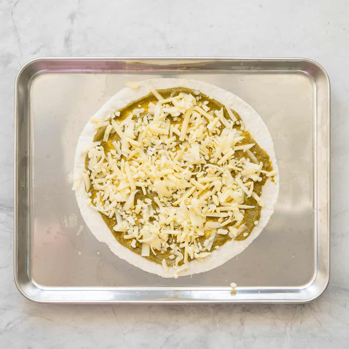 An oven tray resting on the bench with a tortilla spread with vegetable pizza sauce and cheese sprinkled on top of it.
