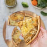 A hand holding a pizza slice topped with veggie pizza sauce above a bench.