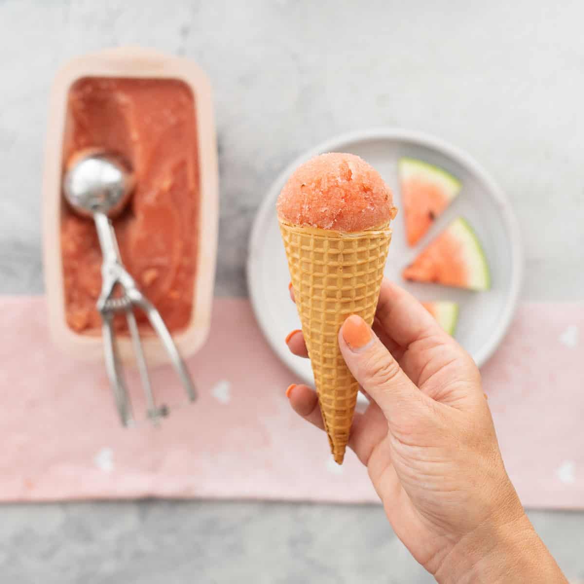 A waffle cone topped with pink sorbet being held above a plate of watermelon slices.