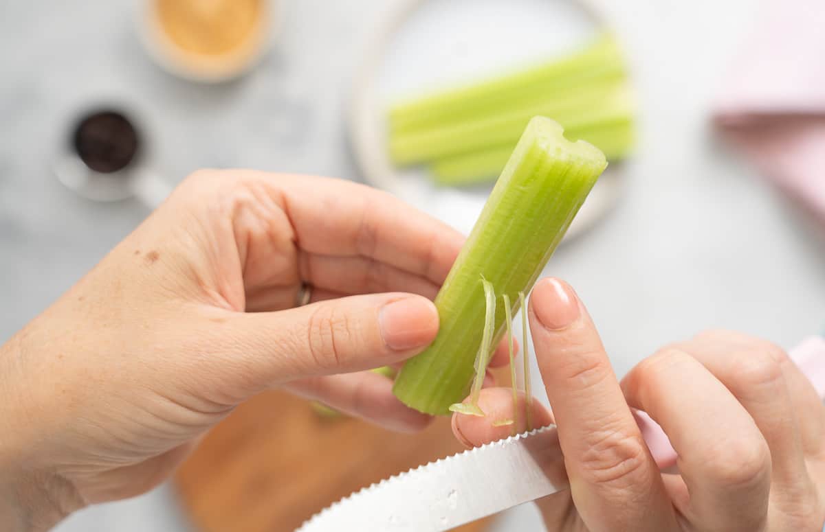 A pair of hands destringing a celery stick with a knife.