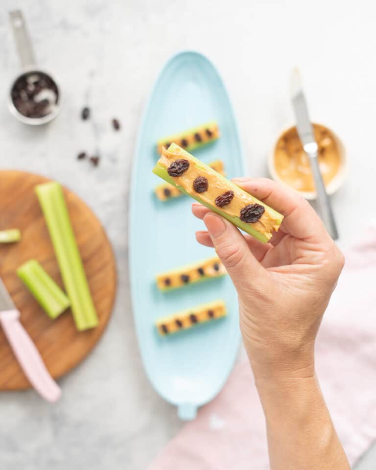 A hand holding a piece of celery filled with peanut butter topped with 4 raisins.