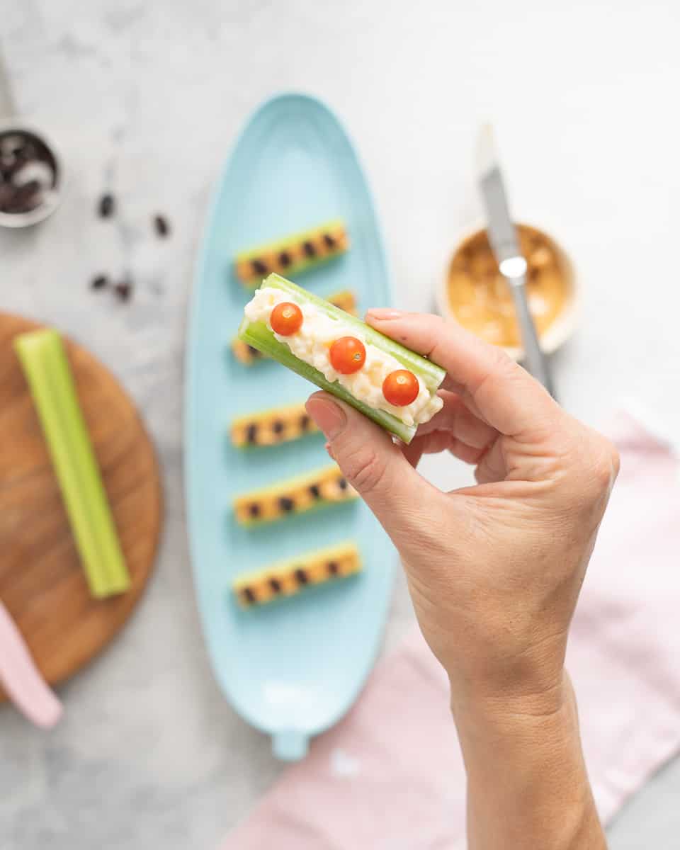 A hand holding a celery stick which is spread with mayonnaise and cheese mix and baby tomatoes on top of it.