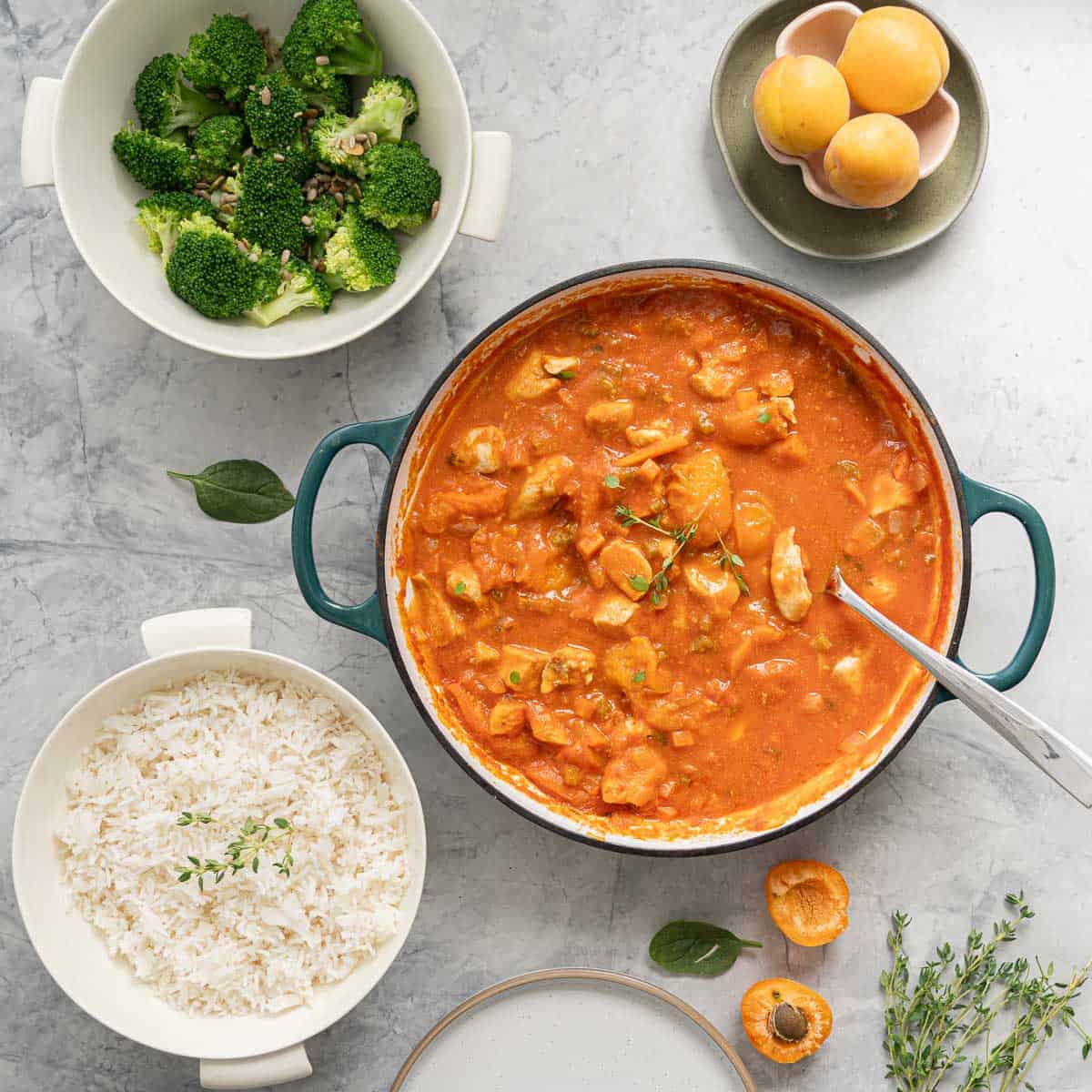A pan full of apricot chicken, a bowl of rice and a bowl of broccoli sitting on the bench alongside fresh apricots and a bunch of time on the bench