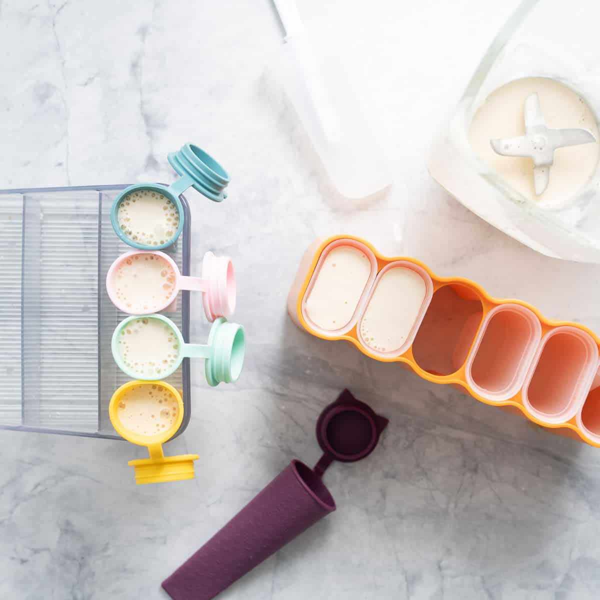 Different shaped and coloured silicon popsicle moulds filled with the banana mixture sitting on the bench next to a blender