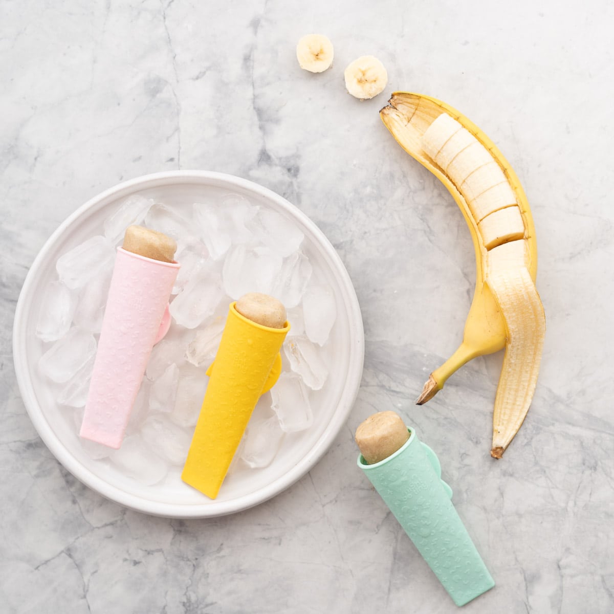 A plate with two banana popsicles resting on top of cubed ice sitting on the bench with one other popsicle laying down beside it.