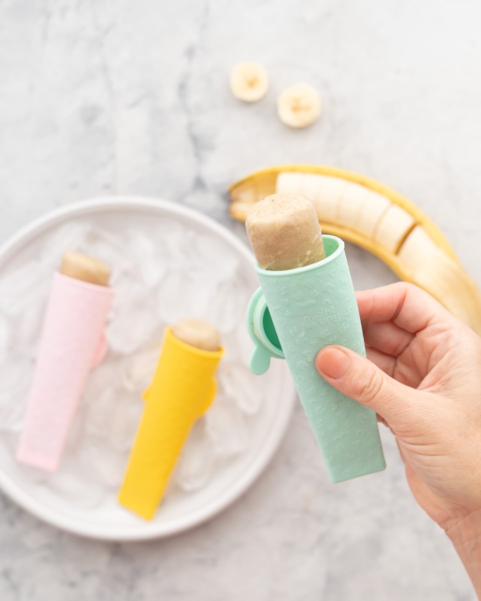 A hand holding a banana popsicle in a silicon mould above two popsicles below which is next to a sliced banana in its skin on the bench