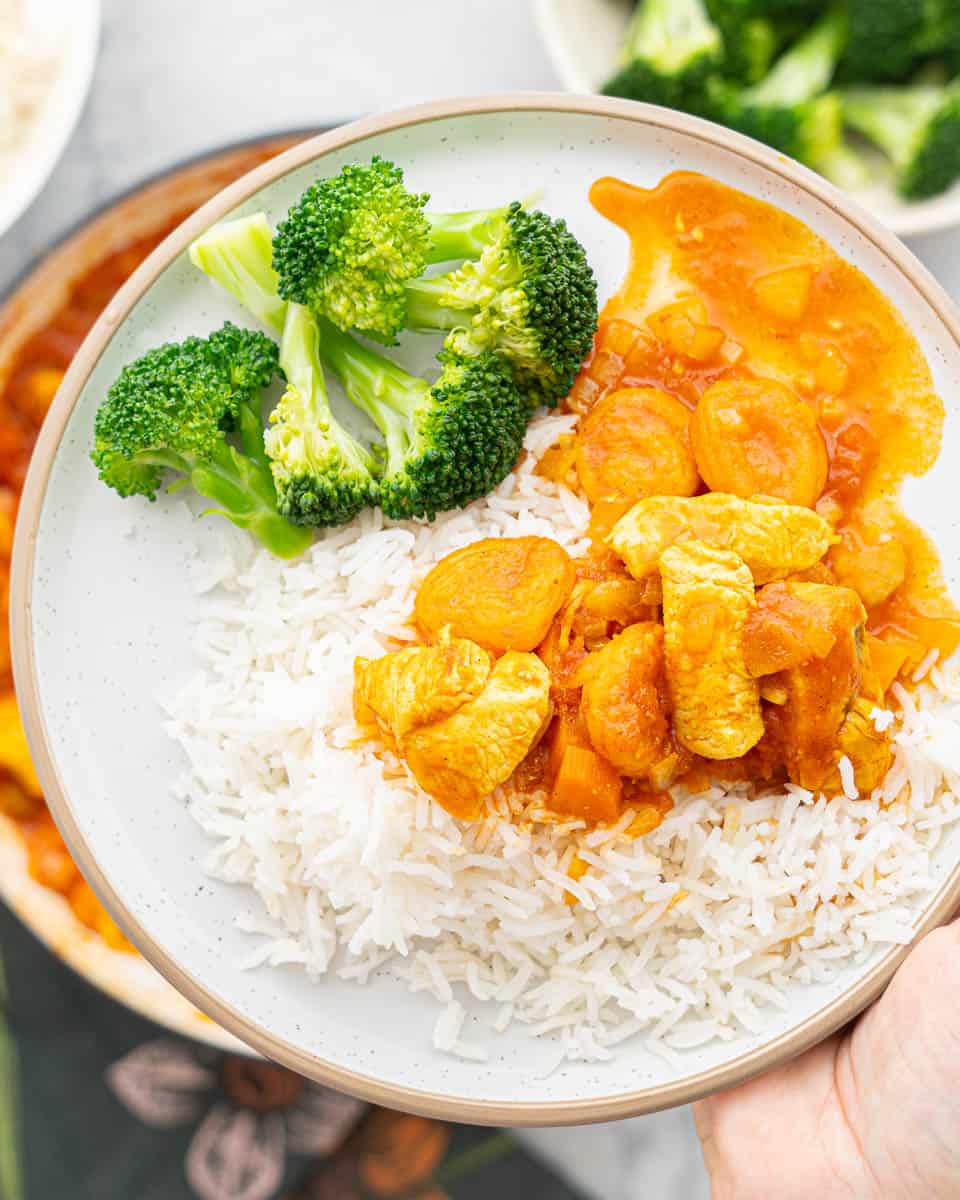 A serving of chicken curry over a bed of rice next to a portion of broccoli on a plate held up above a large pan of curry below it.