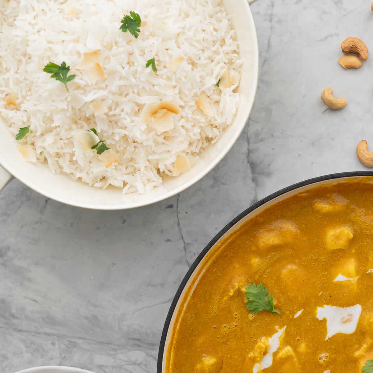 A large bowl of Basmati rice, sprinkled with parsley and a scattering of toasted almonds resting on the bench next to a large pan of chicken curry,