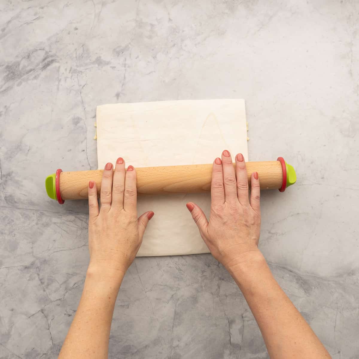 Hands using a rolling pin to press layers of puff pastry and grated cheese.