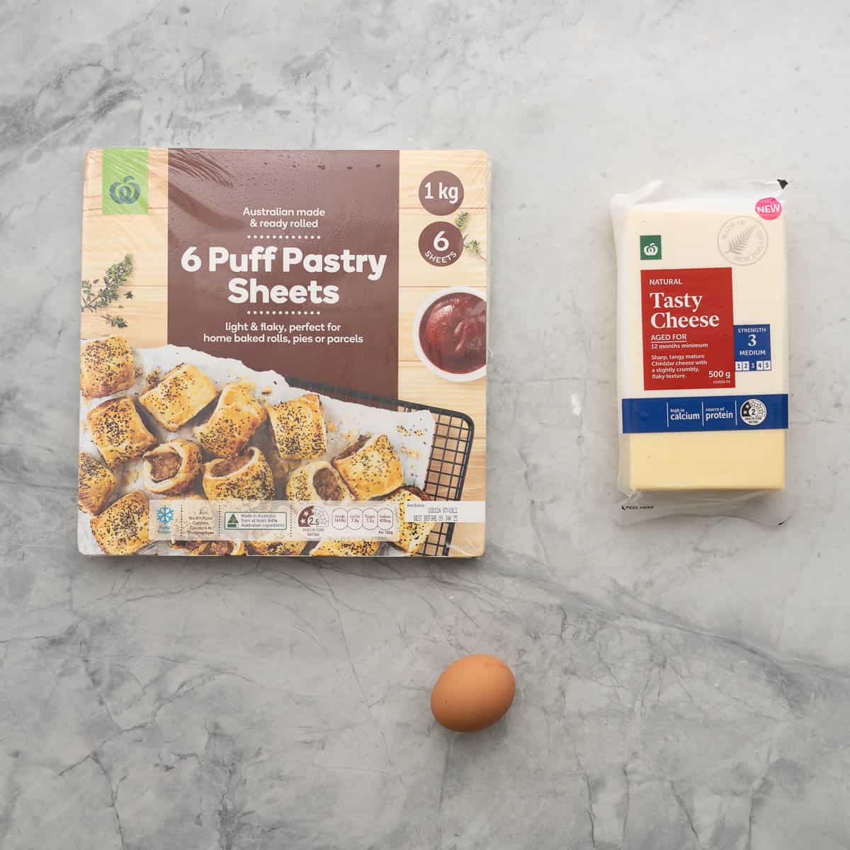 Puff pastry sheets, a block of tasty cheddar cheese and ab egg laid out on a grey marble bench top.