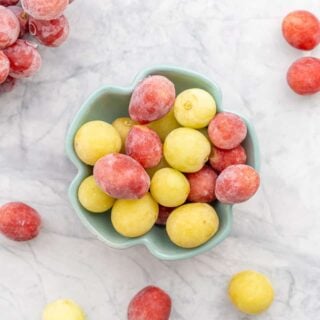 Frozen green and red grapes in a bowl sitting on the bench with some scattered next to it.