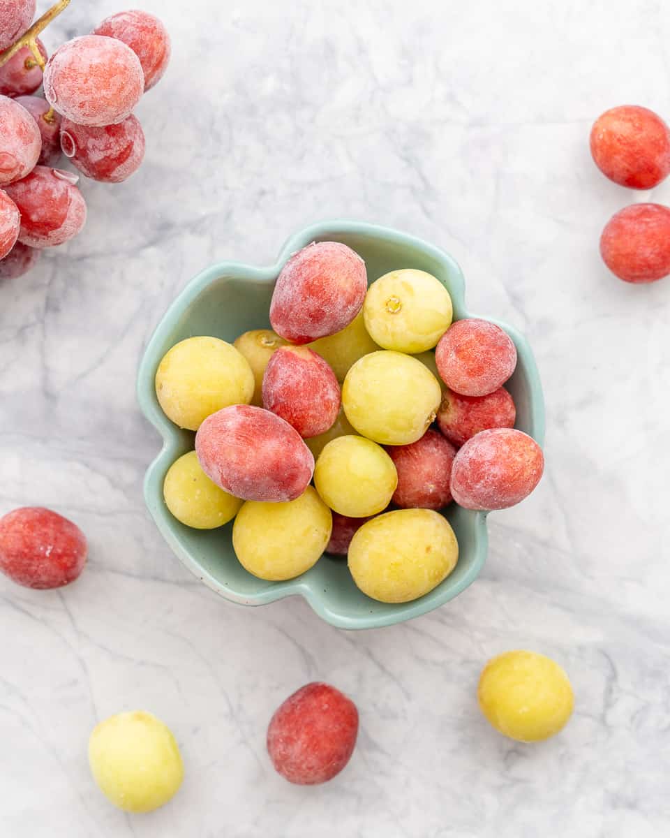 Frozen green and red grapes in a bowl sitting on the bench with some scattered next to it. 
