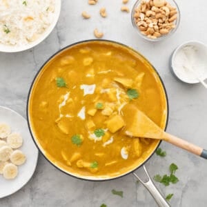 A large pan full of Mango Curry, drizzled with cream and sprinkled with parsley sitting on a bench next to a small bowl of cashew nuts, a ramekin of coconut, a bowl of rice and a plate of sliced bananas.