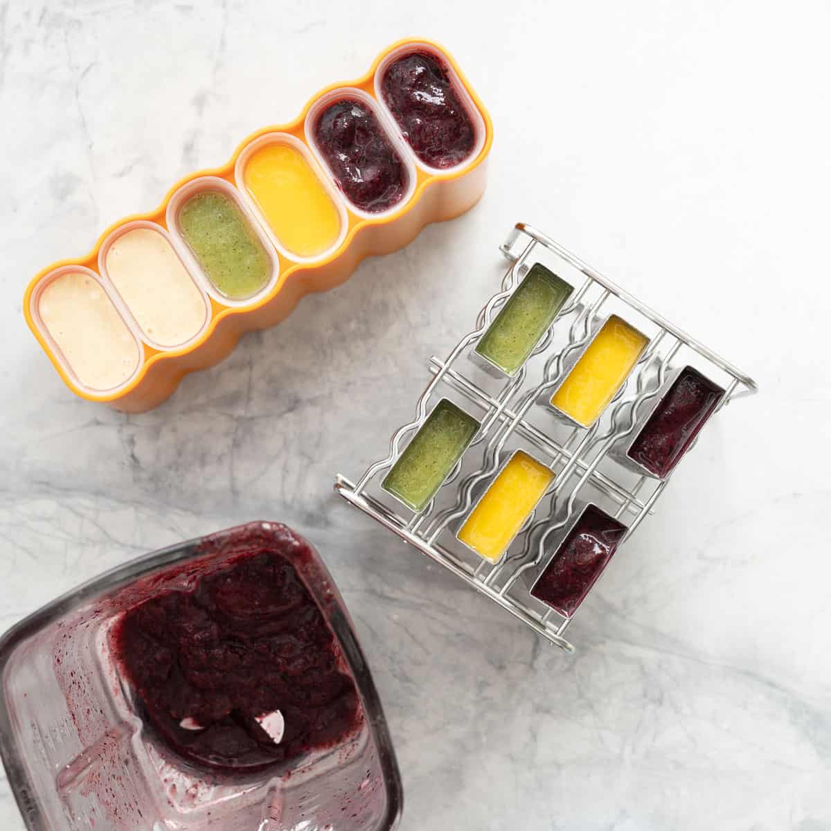 An array of different moulds resting on the bench filled with the different popsicle mixes next to a blender full of the blueberry mixture.