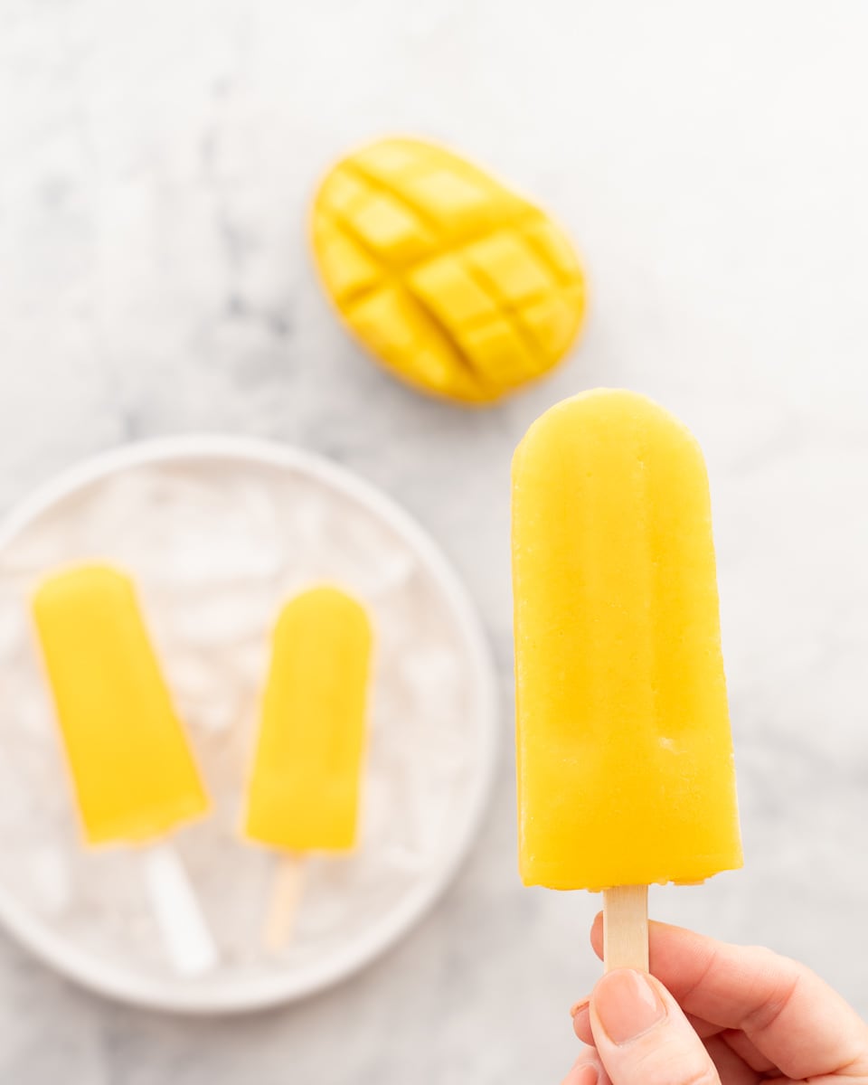 A hand holding a mango popsicle above a plate full of popsicles which is resting on the bench next to a halved mango