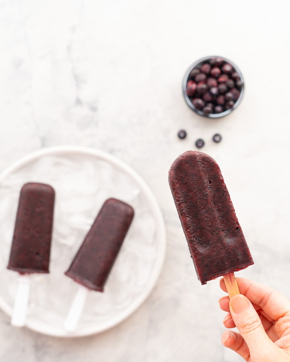 A hand holding a blueberry popsicle next to a plate full of popsicles resting on ice which is next to a small bowl of blueberries.