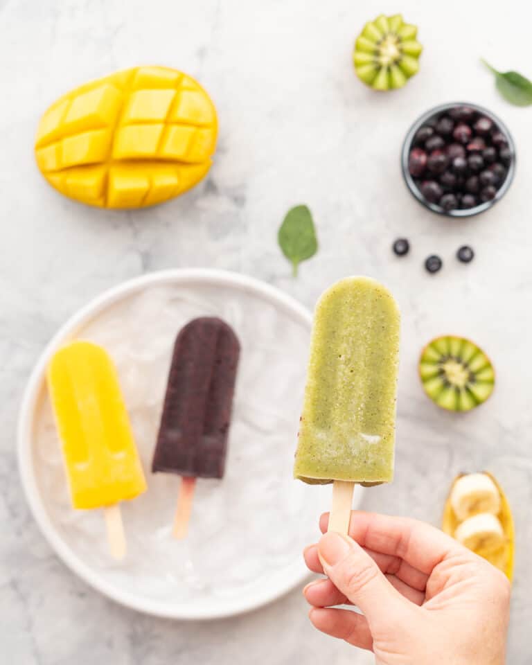 A green kiwi popsicle being held above a plate of brightly coloured fresh fruit and popsicles.