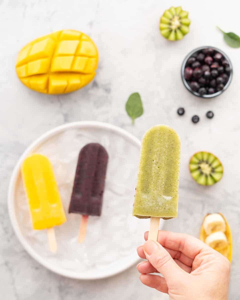 A hand holding a kiwifruit popsicle above a plate which has one each of a mango and blueberry popsicle resting on it. The bench surrounding the plate has got sliced mango, kiwifruit, blueberries and bananas scattered around it.