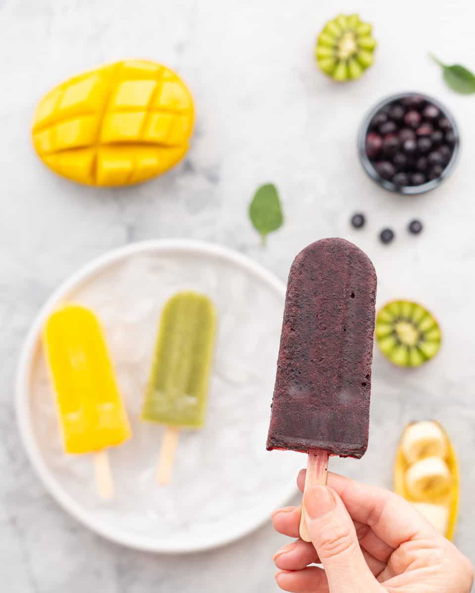 A hand holding a kiwifruit popsicle above a plate which has one each of a mango and blueberry popsicle resting on it. The bench surrounding the plate has got sliced mango, kiwifruit, blueberries and bananas scattered around it.