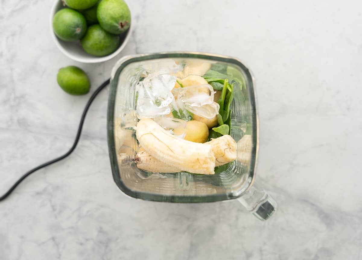Banana, feijoa, spinach leaves and ice cubes in the glass jug of a blender.