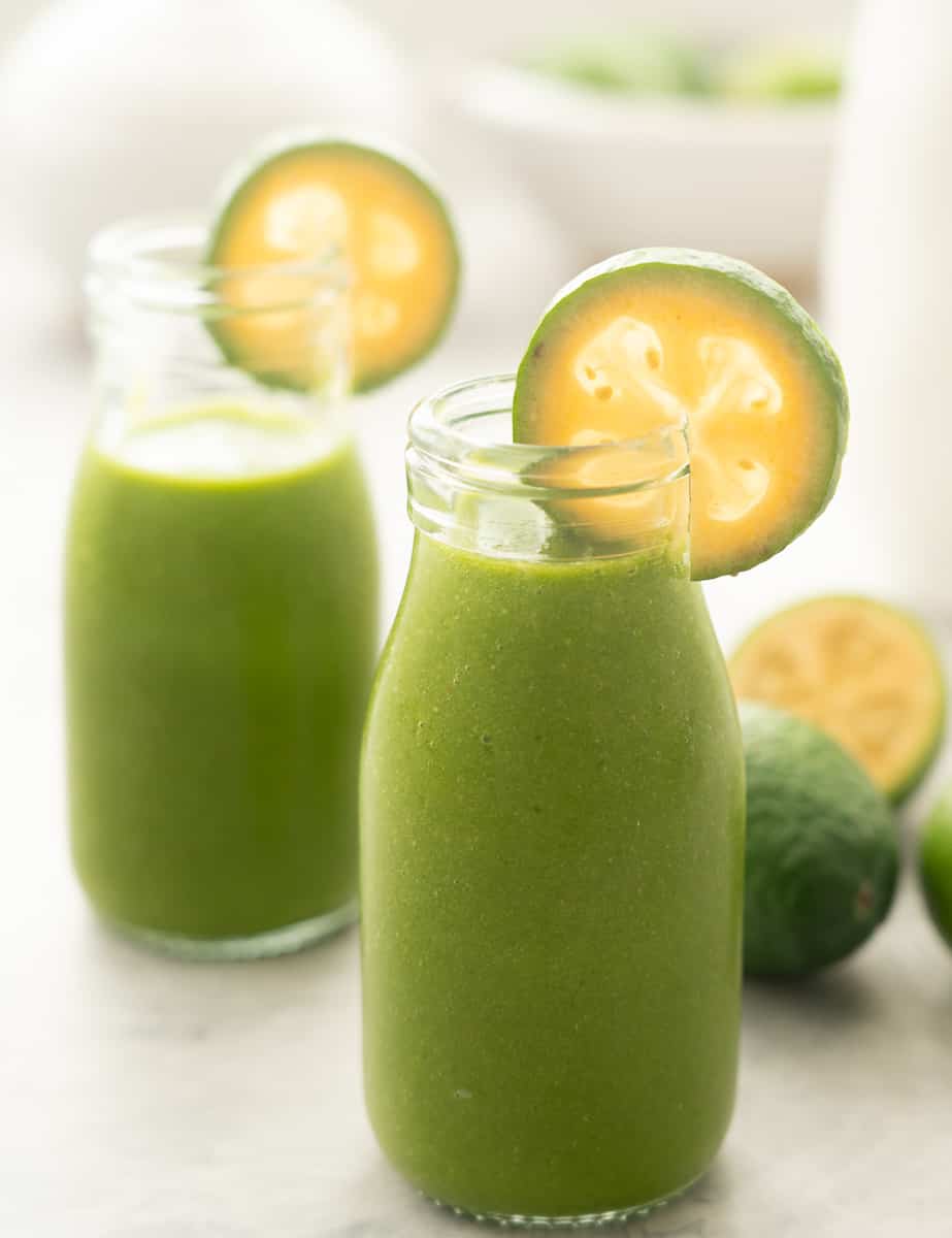 Two small glass bottles filled with green smoothie, the rim of each bottle is garnished with a slice of feijoa.