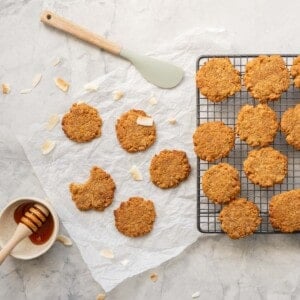 golden cookies on a cooling rack and bench with a dish of golden syrup and coconut chips scattered around them.