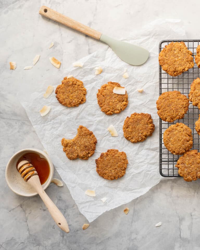 Golden cookies on a cool rack and bench with a dish of golden syrup and coconut chips scattered around them.
