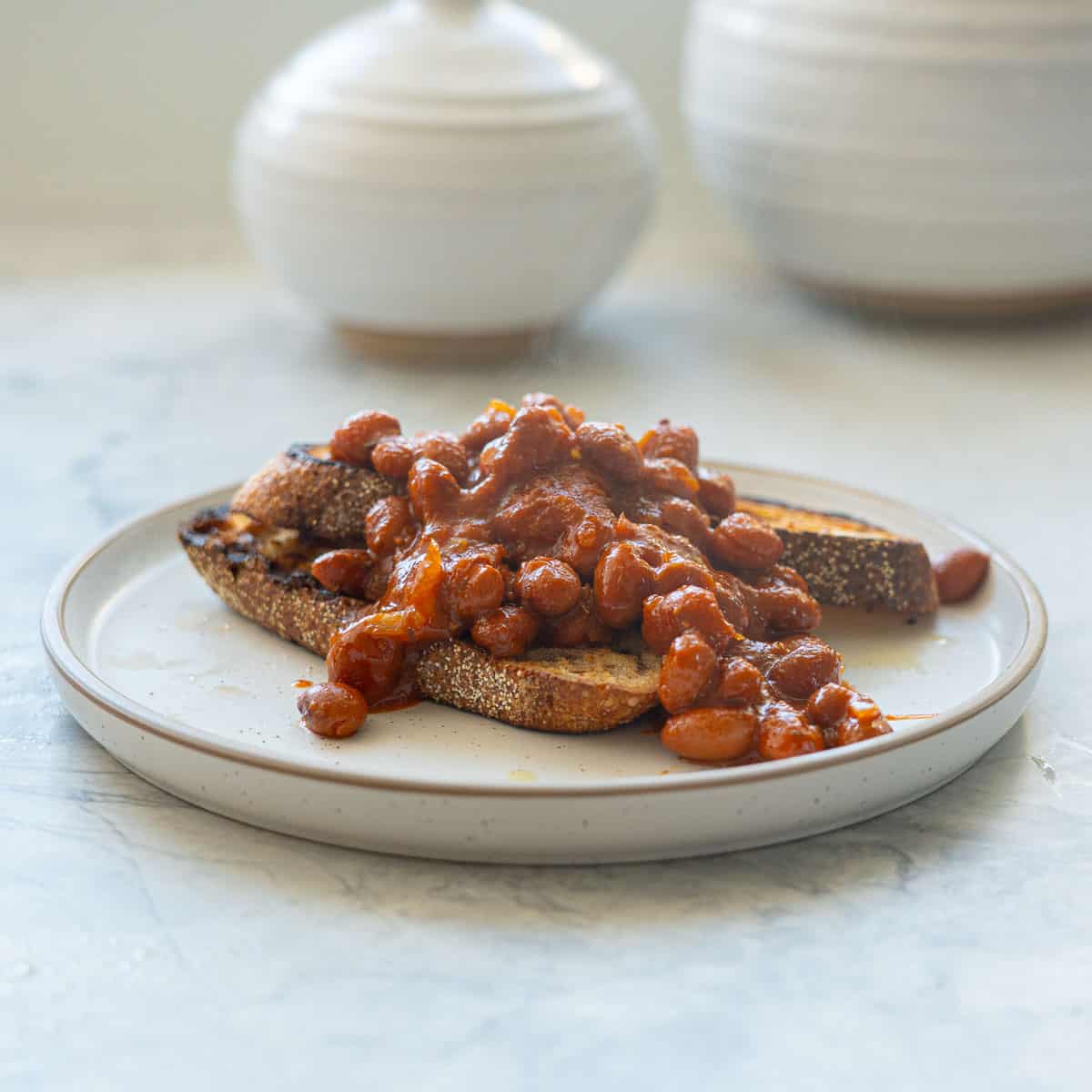 Two slices of rustic toast topped with baked beans on a white plate with brown rim.