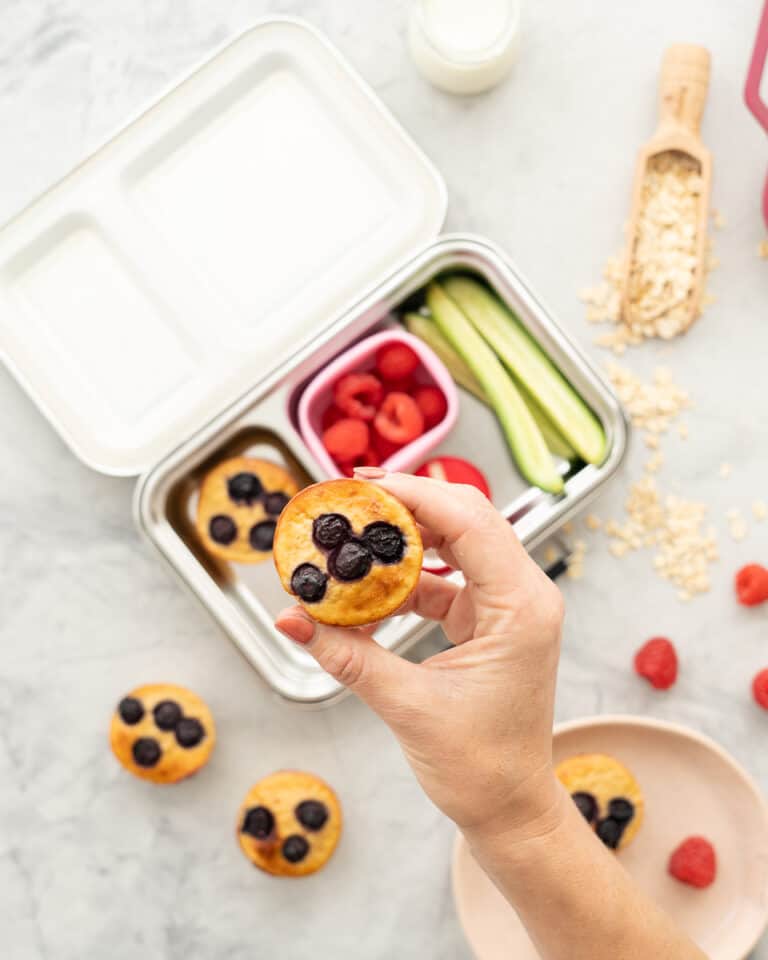 A blueberry topped muffin being held up above a filled stainless steel bento box, raspberries and rolled oats scattered on a bench top.