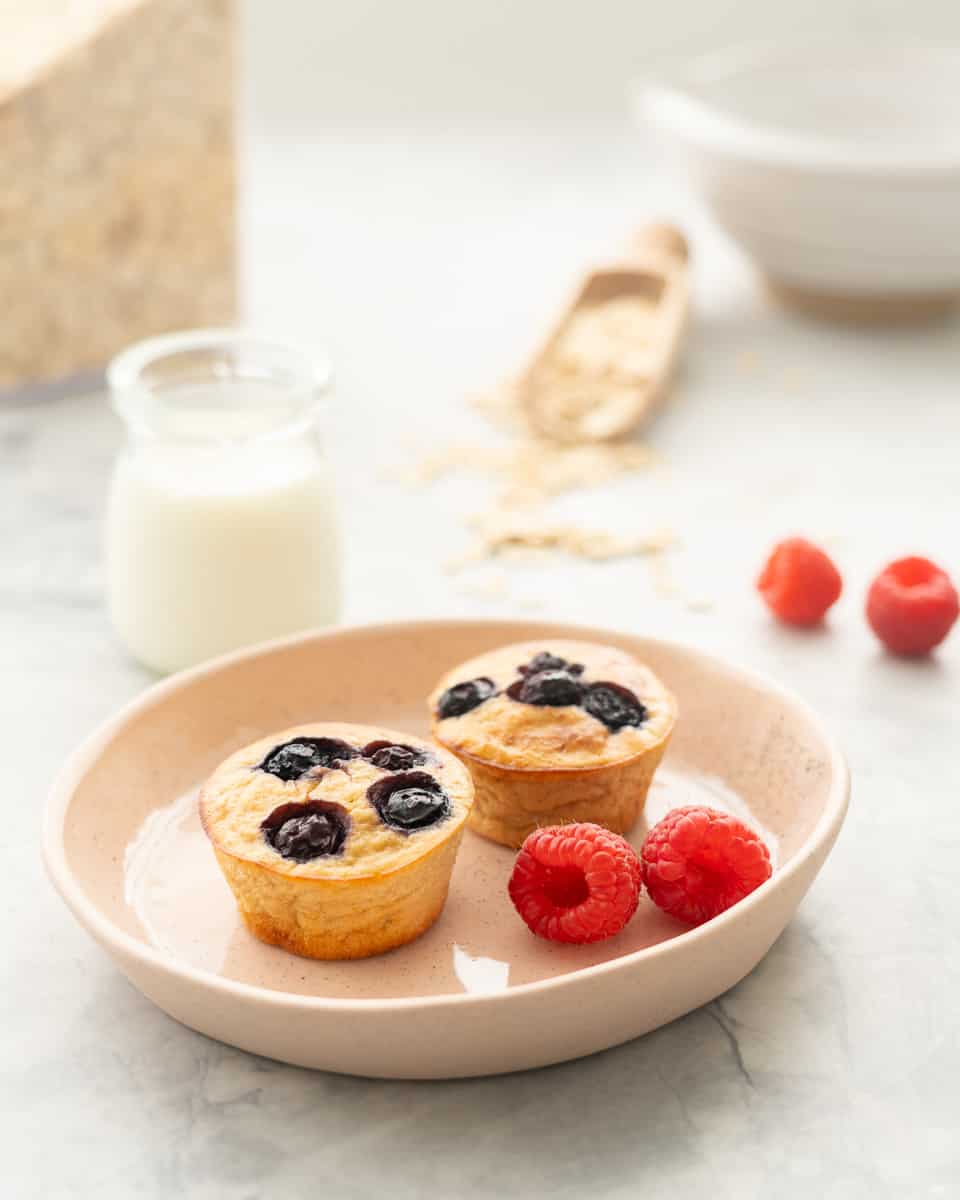 A plate with two blueberry banana oatmeal muffins and fresh raspberries, with glass of milk blurred in back.