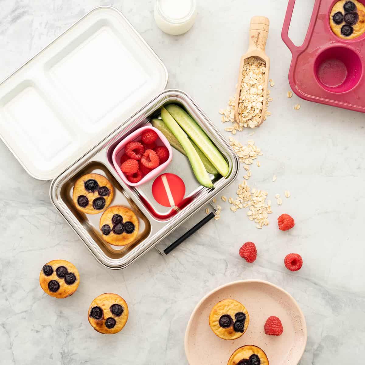A filled stainless steel bento box, with Blueberry banana oatmeal muffins inside, raspberries and rolled oats scattered on a bench top. 